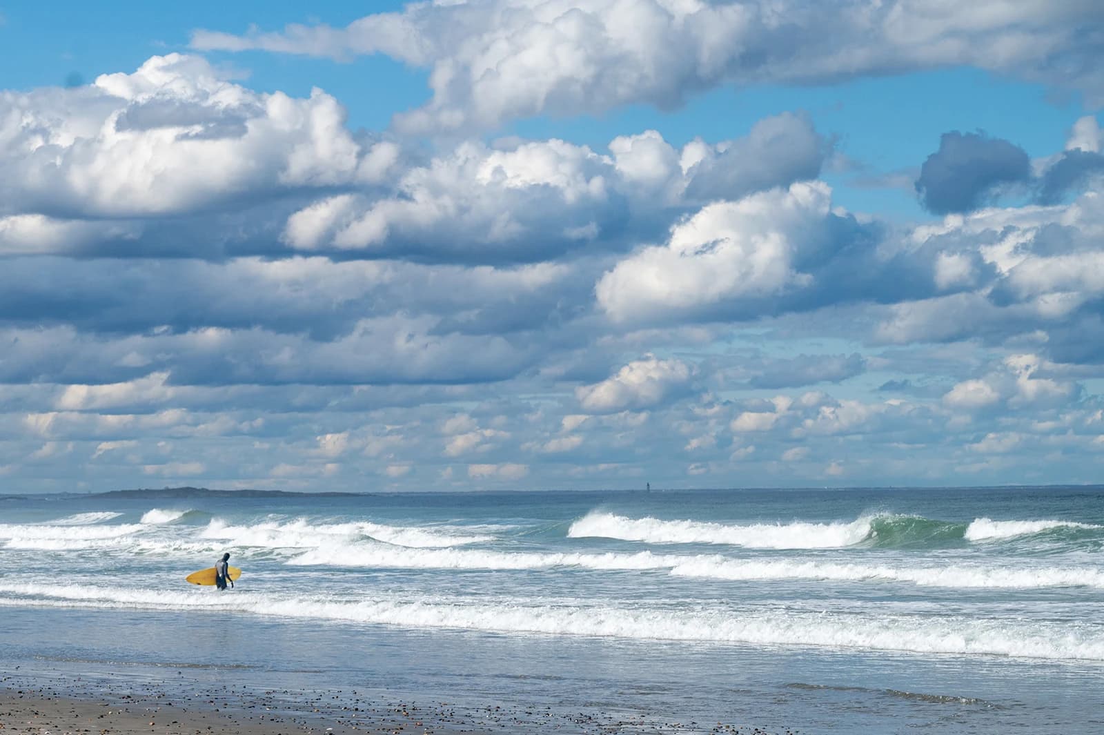 A surfer carrying a yellow board into the waves at Surfside, Hull, under big clouds